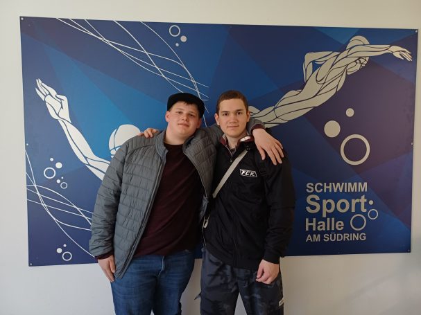 Zwei Männer posieren vor einer blauen Wand mit Schwimm-Motiven und dem Schriftzug "Schwimm Halle".